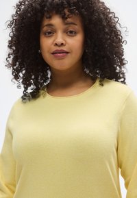 Rumena pulover z zaobljenim izrezom, mehko teksturo in dolgimi rokavi. Ima subtilen meliran vzorec.