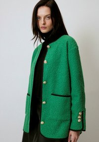 Jachetă verde texturată cu detalii negre, butoni aurii și două buzunare frontale. Designul are o croială lejeră și un decolteu rotund.