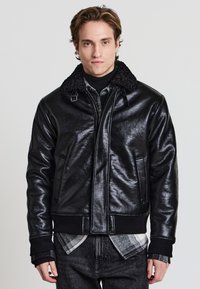 Schwarze Leder-Bomberjacke mit strukturierter Oberfläche, mit einem kuscheligen Kragen, Reißverschluss vorne und gerippten Bündchen. Getragen über einem Rollkragenpullover und einem karierten Hemd.