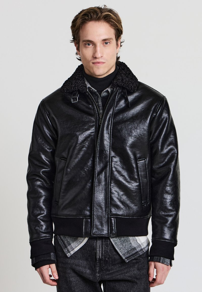 Schwarze Leder-Bomberjacke mit strukturierter Oberfläche, mit einem kuscheligen Kragen, Reißverschluss vorne und gerippten Bündchen. Getragen über einem Rollkragenpullover und einem karierten Hemd.
