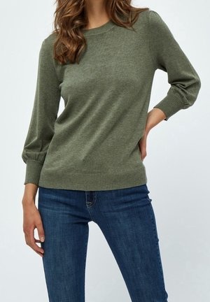 Femme portant un pull vert olive à manches longues avec poignets et ourlet côtelés, associé à un jean bleu foncé, debout avec une main sur la hanche.