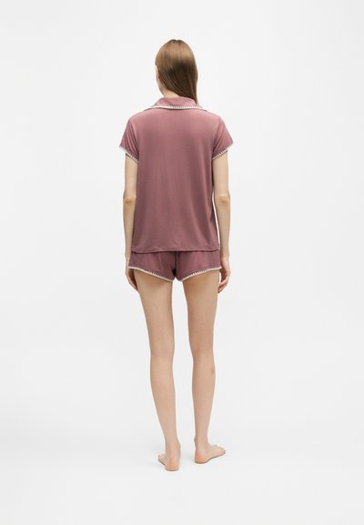 Femme debout pieds nus, de dos, portant un haut mauve à manches courtes et un short assorti avec des bordures blanches aux manches et aux ourlets.