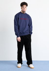 Sudadera azul marino con el logo rojo de "Carhartt", combinada con pantalones de pana negros y zapatillas blancas con suelas de goma, de pie contra un fondo claro.