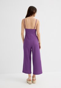 Mono morado de canalé con tirantes finos, piernas anchas hasta la altura de los tobillos y un corpiño ajustado. La tela texturizada aporta profundidad.