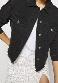 Veste en denim noir au design court, avec un ourlet effiloché et des boutons en argent, superposée à un t-shirt blanc noué et une jupe à motifs.