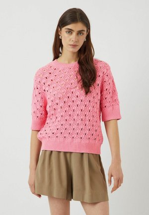 Rosa stickad tröja med ett dekorativt öppet vävmönster, korta ärmar och ribbat fåll, i kombination med ljusbruna lösa shorts.