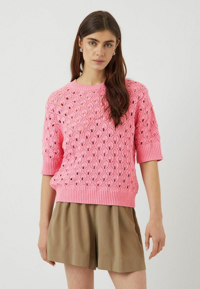 Pull en tricot rose avec un motif de tissage ouvert décoratif, manches courtes et ourlet côtelé, associé à un short ample beige clair.