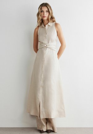 Kvinde med bølget blondt hår iført en ærmeløs beige hørkjole med knapper over matchende bukser med vide ben, stående mod en hvid væg.