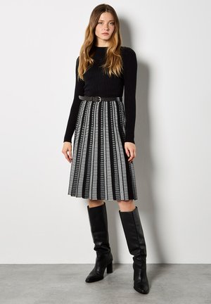 Czarny prążkowany top z długim rękawem w połączeniu z czarno-białą, plisowaną spódnicą. Modelka ma na sobie czarne kozaki do kolan oraz czarny pasek.