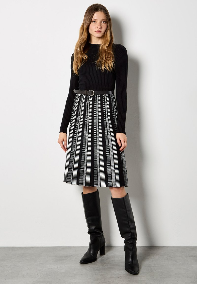 Czarny prążkowany top z długim rękawem w połączeniu z czarno-białą, plisowaną spódnicą. Modelka ma na sobie czarne kozaki do kolan oraz czarny pasek.