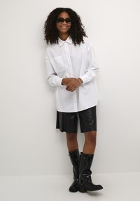 Hvid skjorte med knapper og en lomme, parret med sorte lædershorts og knæhøje sorte støvler. Modellen har krøllet hår og solbriller.