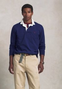 Námornícky dlhý rukáv polo tričko s bielym golierom, béžové nohavice s výšivkami a pruhovaný opasok. Hodinky na zápästí.