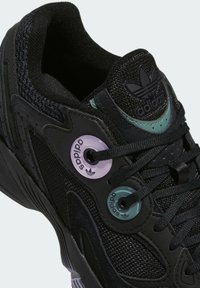 Černé tenisky Adidas s texturovanou černou mesh tkaninou a semišem, s výraznými fialovými a tyrkysovými očky. Logo na jazyku dokončuje design.