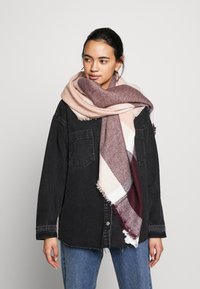 Écharpe en gros tissage de couleur rose pâle, bordeaux et crème avec des bords à franges, drapée sur les épaules et assortie à une veste noire.