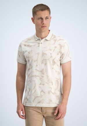 Man draagt een beige polo met patroon en kaki broek, staand tegen een effen lichte achtergrond, kijkend naar de zijkant.
