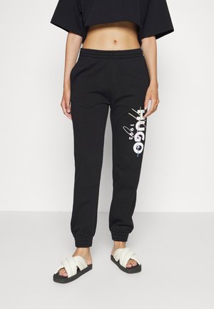 Persoon draagt zwarte joggingbroek met witte "HUGO 1993"-print op het linkerbeen, zwart cropped top en witte gevoerde slidesandalen.