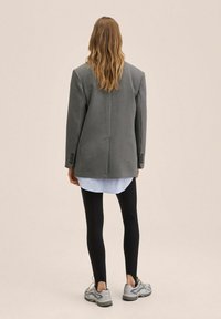 Przesadny żakiet w wzór choćhaka w odcieniach szarości, noszony na niebieskiej koszuli, w połączeniu z czarnymi legginsami i szarymi trampkami.