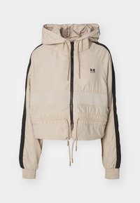 Beskaaret beige jakke med et sort sidesektion, justerbar snørrebånds hætte, elastiske manchetter og lynlås foran. Har logo på brystet.