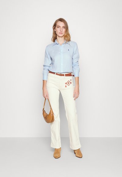 Hellblaues Hemd mit Knopfleiste, cremefarbene Schlaghose mit gestickten Details, brauer geflochtener Gürtel und hellbraune Stiefeletten mit Absatz; hält eine braune Handtasche.