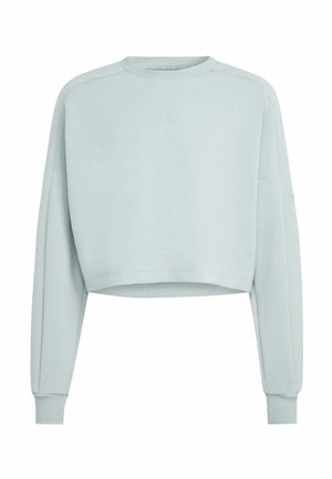 Joven chica con cabello largo lleva una sudadera azul claro de gran tamaño y leggings a juego, de pie frente a un fondo gris liso.