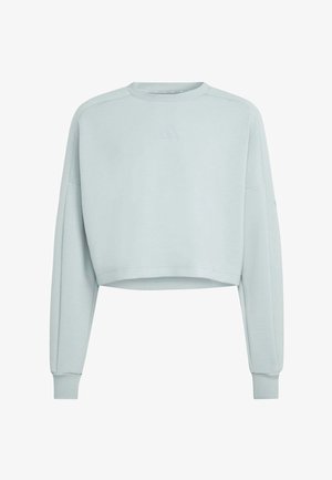 Joven chica con cabello largo lleva una sudadera azul claro de gran tamaño y leggings a juego, de pie frente a un fondo gris liso.