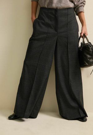 Person trägt weite dunkel karierte Hosen, braunen Pullover, schwarze Schuhe und hält eine schwarze Ledertasche, steht vor einem beigen Hintergrund.