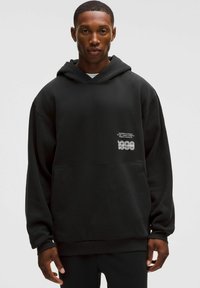 Czarna, oversize bluza z kapturem wykonana z miękkiego materiału, z kieszenią z przodu i białymi nadrukami po lewej stronie. Prosty, uliczny styl.