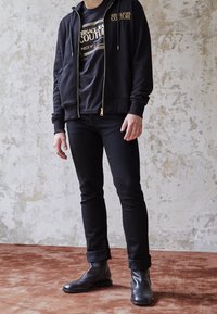 Sudadera negra con letras doradas, camiseta negra debajo, jeans negros ajustados y botas de cuero negra. Atuendo urbano casual sobre un fondo texturizado.
