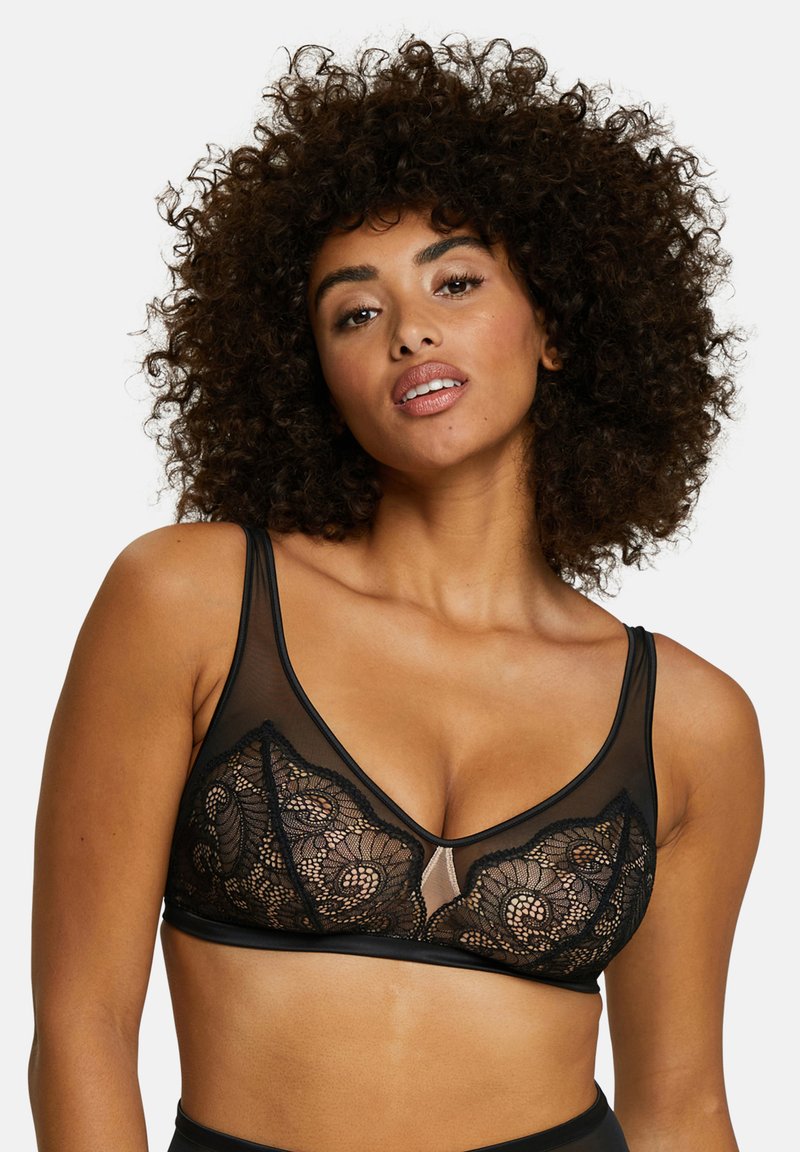 Vrouw met krullend haar die een zwarte kanten bralette met doorzichtige panelen draagt, poserend tegen een effen witte achtergrond.