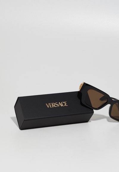 Mustat aurinkolasit ruskeilla linsseillä, kulmikas muotoilu, kultaiset yksityiskohdat temppeissä, asetettu mustan laatikon viereen, jossa on Versace-logo.