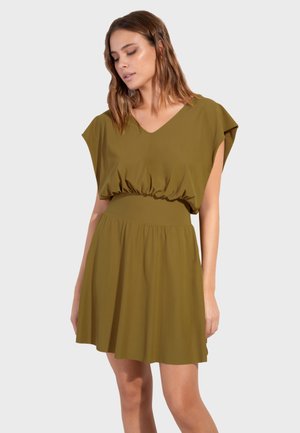 Kobieta ubrana w oliwkowozieloną sukienkę bez rękawów z marszczonym pasem i dekoltem w serek, stojąca na tle jasnoszarej, jednolitej ściany.