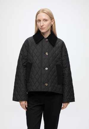 Femme aux cheveux blonds et raides portant une veste matelassée noire avec de gros boutons et un col noir, debout devant un fond blanc uni.