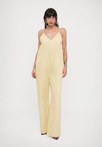 Κίτρινη ολόσωμη φόρμα με βαθύ V-λαιμόκοψη, λεπτές ρυθμιζόμενες τιράντες και φαρδιά εφαρμογή. Διαθέτει μπροστινό κούμπωμα με κουμπιά και φαρδιά γραμμή στο κάτω μέρος.