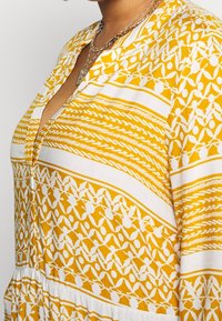 Nära bild av en person som bär ett gult och vitt mönstrat plagg med V-ringning och lager av guldhalsband.