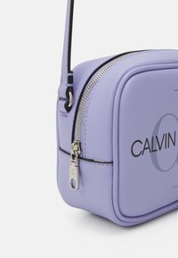 Levandulová syntetická kabelka přes rameno se strukturovaným tvarem, stříbrným zipem a raženým logem "CALVIN" na přední straně. Černé doplňky na řemínku.
