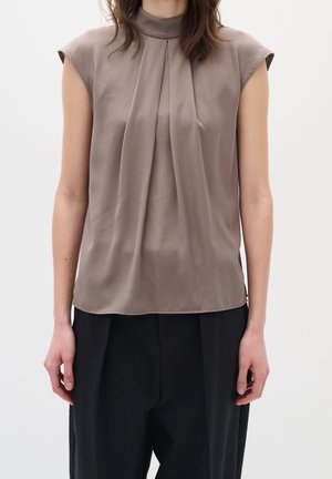 Femme portant un chemisier sans manches taupe avec des plis à l'avant et un col montant, associé à un pantalon noir, debout devant un fond blanc uni.