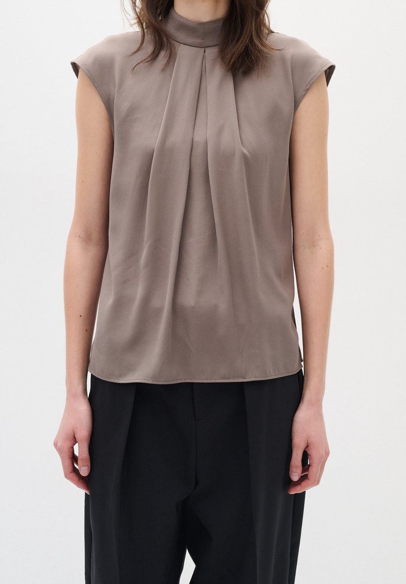 Femme portant un chemisier sans manches taupe avec des plis à l'avant et un col montant, associé à un pantalon noir, debout devant un fond blanc uni.