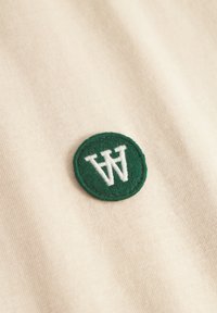 Beige stof med en tekstureret overflade har et cirkulært grønt broderet mærke, der viser et hvidt "W" logo.