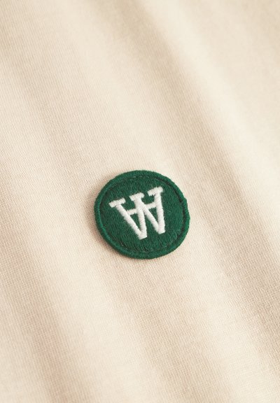 Beige Stoff mit strukturiertem Oberflächenbild zeigt ein kreisförmiges grünes gesticktes Emblem mit einem weißen "W"-Logo.