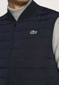 Tmavě modrá péřová vesta s horizontálním prošíváním, zapínáním na zip a černým žebrovaným límcem. Na hrudi je zeleně vyšívané logo krokodýla.