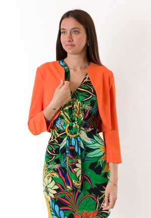 Giacca cropped arancione brillante sopra un vestito a fantasia con design floreali verdi, blu e marroni, stretto in vita con una cintura circolare.
