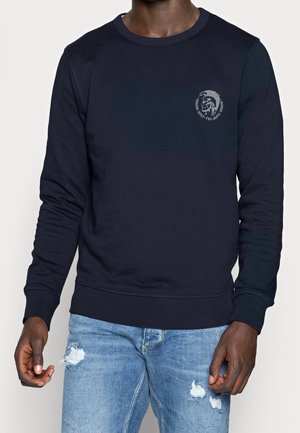 Homme portant un sweat-shirt bleu marine à col rond avec un petit logo sur la poitrine et un jean bleu clair déchiré, debout devant un fond uni.