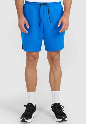 Mężczyzna ubrany w jasnoniebieskie sportowe spodenki z czarnym sznurkiem, białe skarpety sportowe i czarne buty do biegania, stojący na szarej podłodze.