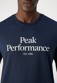 Laivastonsininen puuvillapaita, jossa on valkoiset tekstit "Peak Performance" ja "Est. 1986". Pyöreä kaulus, sileä pinta ilman näkyviä kuvioita.