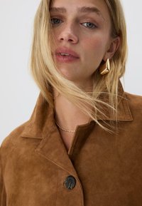 Blond kvinna med löst hår bär en brun mockajacka, ett guld snurrat kedjehalsband och stora, glänsande geometriska örhängen i guld.