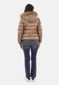 Giacca a vento imbottita color beige chiaro con cappuccio foderato in pelliccia, design trapuntato e silhouette aderente. Abbinata a jeans blu e scarpe da ginnastica bianche.