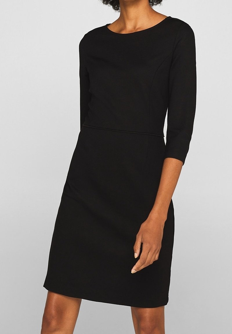 Femme portant une robe noire longueur genoux avec des manches trois-quarts et un col rond, debout devant un fond gris clair uni.