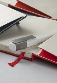 Penna bianca con clip grigia su un quaderno aperto con pagine crema e segnalibro rosso; cartelle blu e rosse sullo sfondo, con copertine semplici.