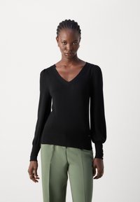 Schwarzer Strickpullover mit V-Ausschnitt, voluminösen langen Ärmeln und geripptem Saum. Kombiniert mit hellgrünen Hosen im entspannten Schnitt.