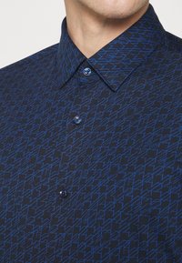 Niebieska koszula w geometryczny wzór na ciemnym tle, z kołnierzykiem na guziki i pasującymi niebieskimi guzikami.